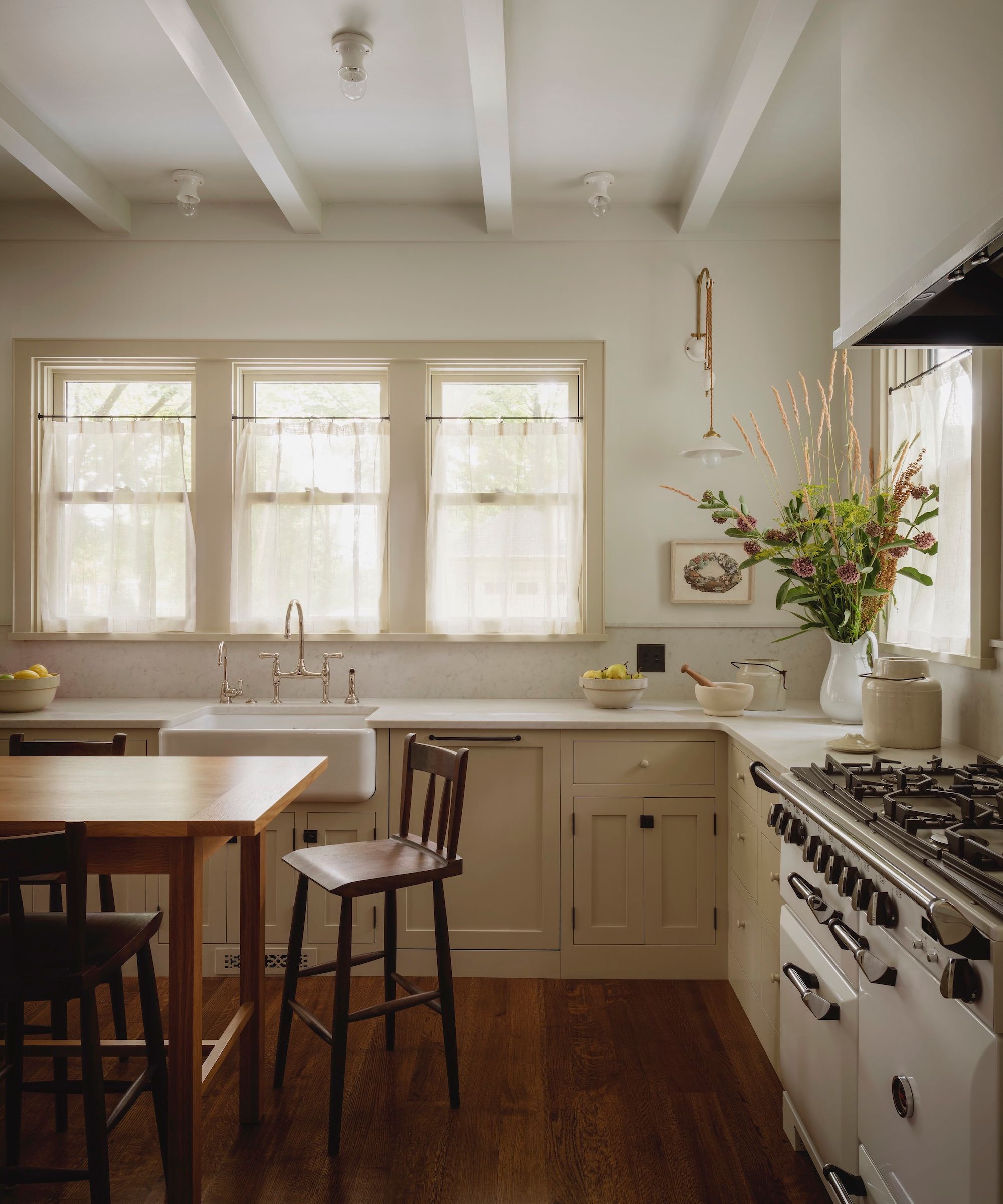 A modern rustic kitchen with cream-colored cabinets, a row of three windows with sheer cafe curtains, warm wood flooring, white walls with ceiling beams, and a wooden dining table with bar chairs.