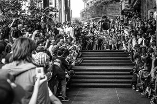 Black and white image of Go Skate Day, Vancouver, 2009, crowd of people watching and taking photographs, skateboarder in mid air suspended over stone steps, surrounding landscape of trees and buildings in the back drop