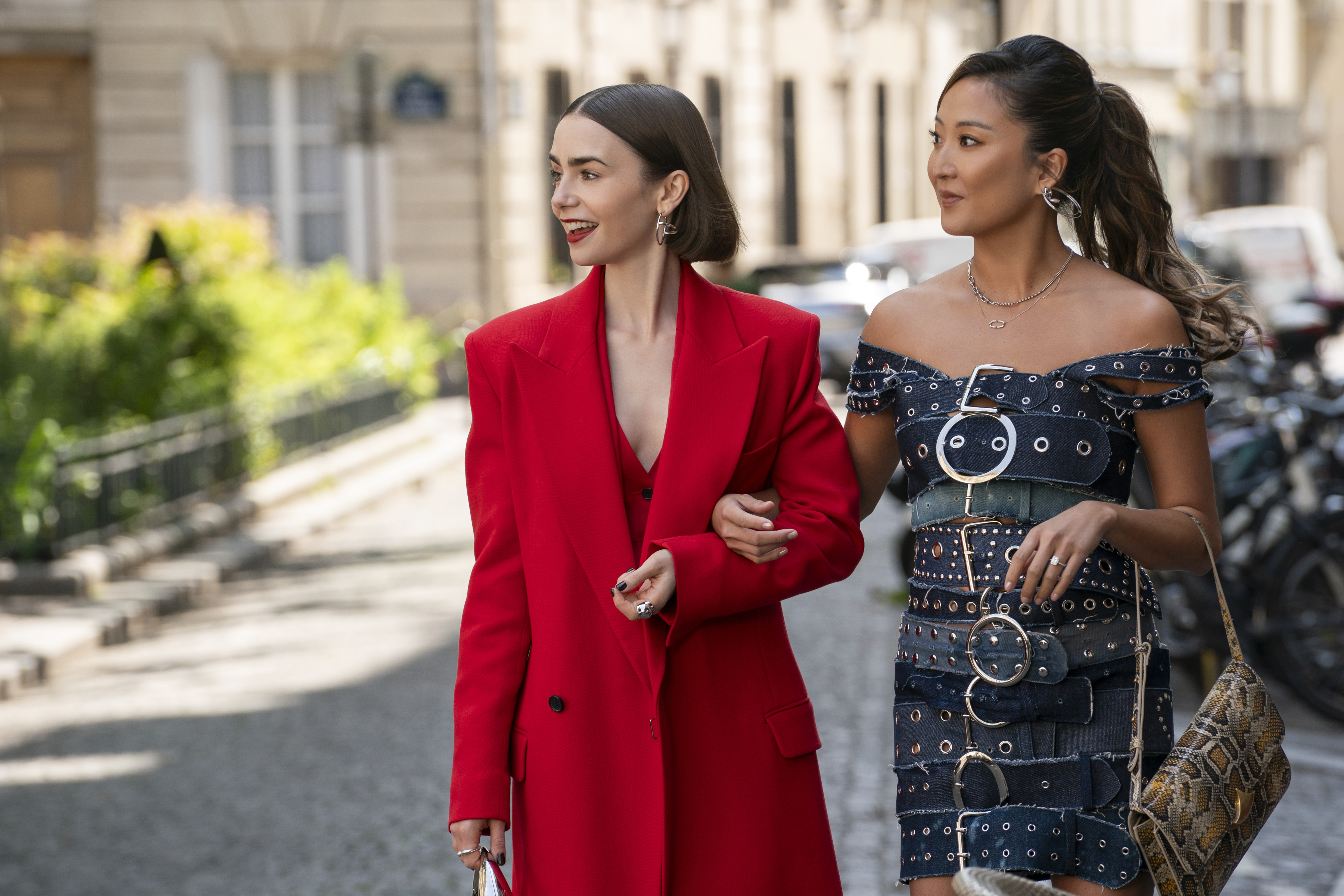 lily collins as emily in a red suit and ashley park as mindy in a belted dress walking arm in arm through paris in emily in paris season 5