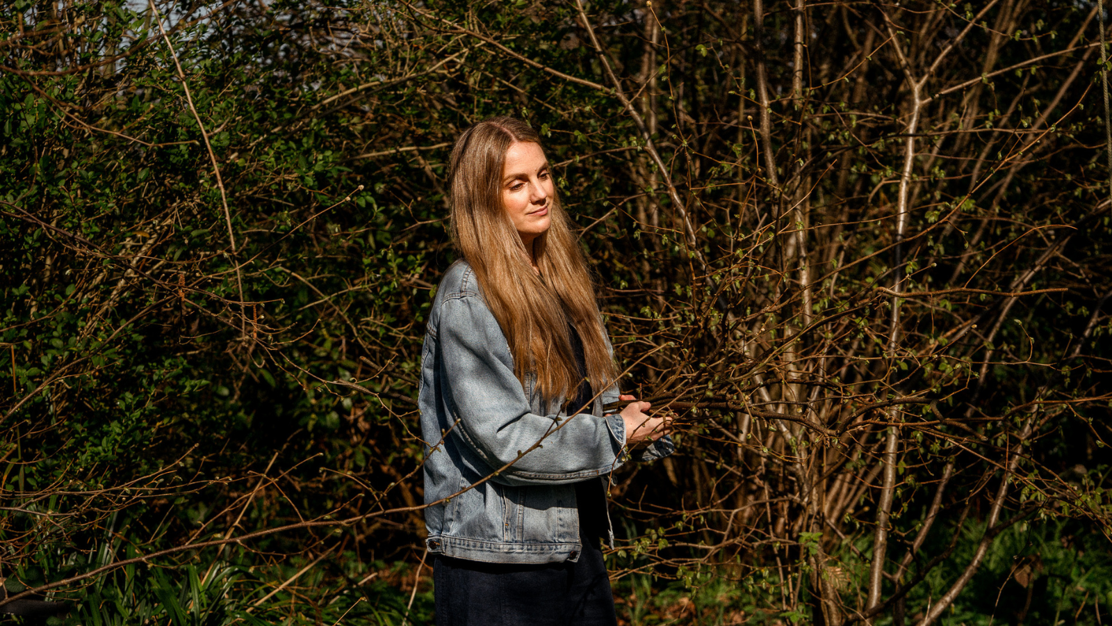Woman in blue denim jacket carrying cut hazel branches through woodland garden