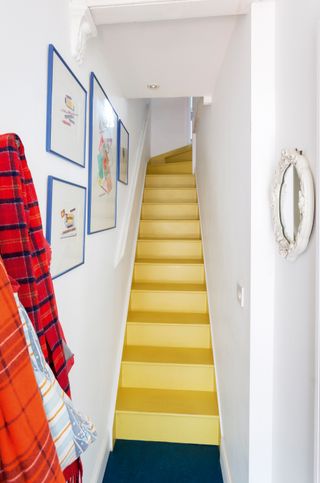 yellow narrow staircase in a white hallway, with blue artwork on the walls and scarves hanging up on the wall