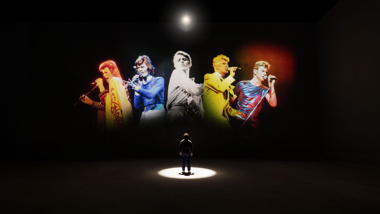 A person stands spotlit in a dark room in front of projected images of David Bowie in five different guises for the exhibition David Bowie: You're Not Alone at Lightroom, London.