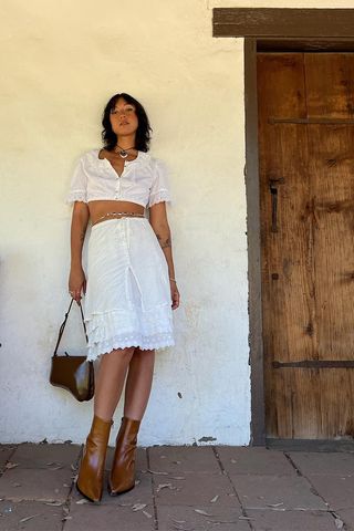 a photo of a woman wearing brown heeled ankle-boots in summer with white lace skirt and matching cropped top with a choker necklace