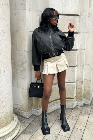 a photo of a woman wearing black heeled ankle-boots in summer with a khaki pleated mini skirt and leather bomber jacket