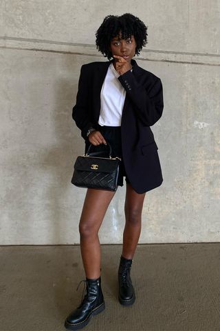 a photo of a woman wearing black ankle-boots in summer with black shorts, a white t-shirt, and a black blazer and a quilted handbag