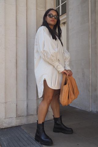 a photo of a woman wearing black ankle-boots in summer with white button-down shirt dress and tan handbag and big sunglasses