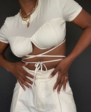 a picture of a white outfit idea for summer with a woman wearing a white tie-up crop top with white trousers and layered chains
