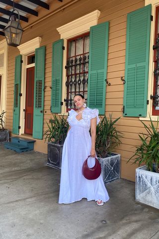 a picture of a white outfit idea for summer with a woman wearing beaded earrings, a white ruffled maxi dress, white sandals, and a red bag