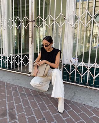 A woman wearing white jeans, a black top, white boots, and a beige tote bag.