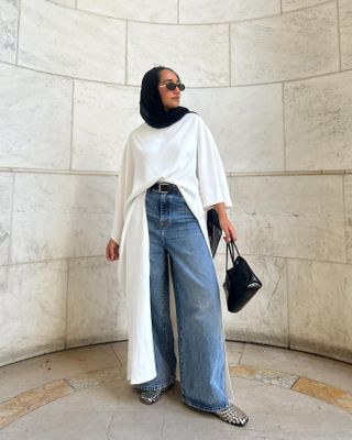 Yusra wearing wide-leg jeans, studded ballet flats, a long white top, and a black handbag.