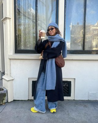 Amaka Hamelijnck wearing baggy jeans, yellow sneakers, a long coat, a blue scarf, blue hat, and brown crossbody bag.