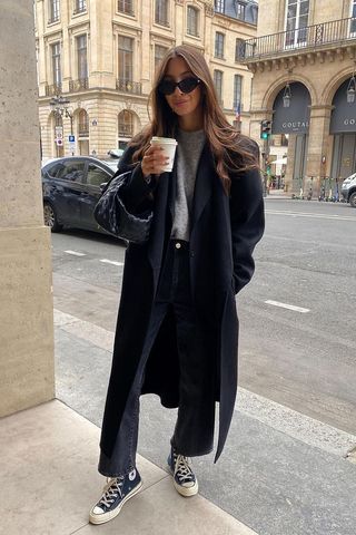 a photo showing how to wear wide jeans with a woman wearing black wide leg jeans with a gray sweater, black coat, converse sneakers, and a woven bag