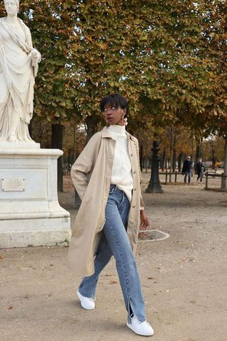 a photo showing how to wear wide jeans with a woman wearing wide leg jeans with a white turtleneck, white sneakers, and a trench coat