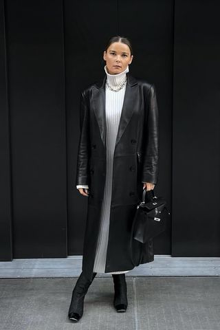 a photo showing how to wear dresses in the winter with a woman wearing a black long leather coat over a white turtleneck dress with black knee-high boots