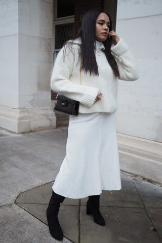 a photo showing how to wear dresses in the winter with a woman wearing a white turtleneck dress under a matching zip-up jumper styled with black knee high boots and a black shoulder bag