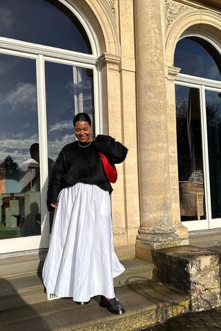 a photo showing how to wear dresses in the winter with a woman wearing a black crewneck sweater over a white maxi dress with black loafers and a red satin bag