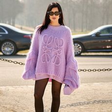 A woman wears sheer tights with a purple Loewe sweater, a pink long-sleeve shirt, a pink Loewe puzzle bag, and black sunglasses.