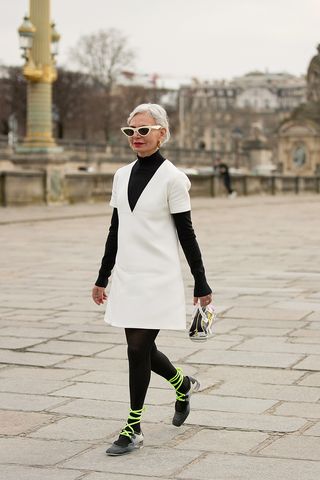 Grece Ghanem wears black sheer tights, a black long-sleeve, white minidress, white sunglasses, and lace-up sneakers.