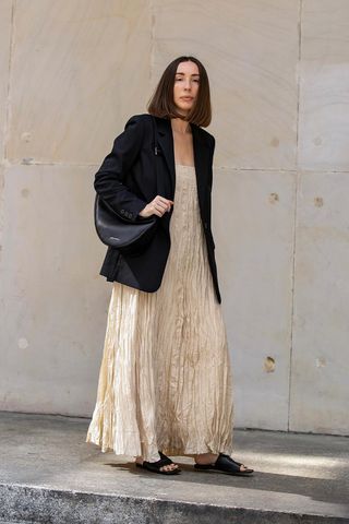 a photo of a woman's outfit with flat black slide sandals with a maxi creme crinkled dress and a black blazer and black shoulder bag