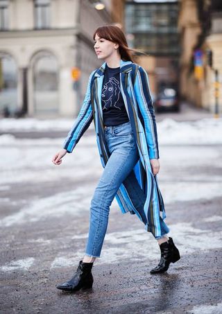 A woman wearing a blue striped long coat, cropped jeans, black ankle boots, and a blue graphic t-shirt.