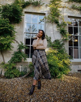 A woman wearing a plaid maxi skirt, a brown turtleneck, and black ankle boots.