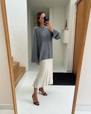 A woman wears a gray cable-knit sweater, a white slip skirt, and black heels.