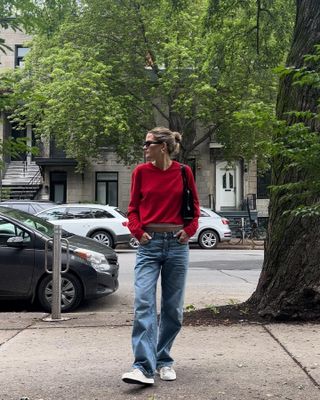 Eliza wears a red sweater, baggy jeans, and white sneakers.