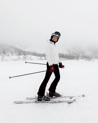 A woman wears a white ski jacket, black ski pants, and burgundy gloves.