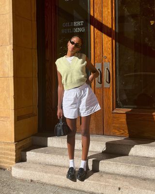 influencer Claire Most poses on steps outside of a building wearing black oval sunglasses, a yellow sweater vest, white cotton pull-on shorts, white ankle socks, and black loafers