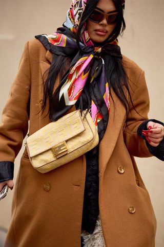 A woman wears a butter yellow Fendi Baguette bag with a printed silk head scarf, tan wool coat, and red sunglasses.