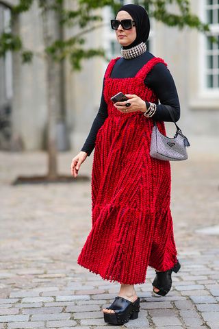 A woman wears the Prada Tessuto Re-Edition Shoulder Bag in a studded version with a red maxi dress, black heels, black long-sleeve shirts, studded choker, and black sunglasses.