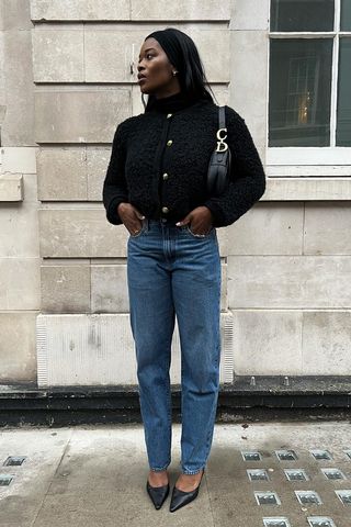 a photo showing what shoes to wear with bootcut jeans, specifically a woman wearing a black tweed jacket with a black dior bag, dark wash bootcut jeans, and pointed black mules