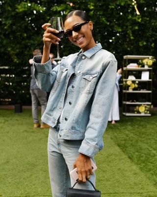 Influencerin Tylynn Nguyen posiert beim Anstoßen mit einem Glas Wein und trägt eine Cat-Eye-Sonnenbrille, eine Jeansjacke und Jeans.