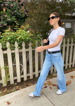 Judith wears blue wide-leg jeans, white adidas samba sneakers, a black belt, a white t-shirt, black sunglasses, and a black handbag.