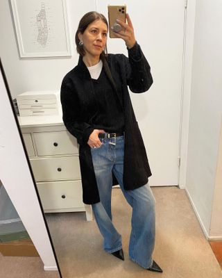 Judith wears blue wide-leg jeans, a black belt, black pointed-toe shoes, a black sweater, white t-shirt, and black long coat.