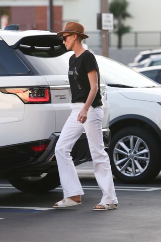 Charlize Theron wearing black graphic t-shirt, white jeans, and white birkenstocks in parking lot