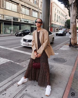 A woman wears a tan blazer, floral maxi dress, white sneakers and a burgundy bag.