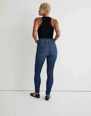 Woman wearing Madewell Curvy 10-Inch High-Rise Skinny Jeans with black tank top and black flats against white background, back turned to camera