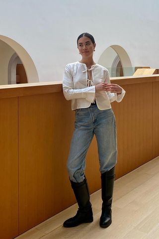 riding boot outfit shown in a photo of a woman in an art museum wearing dainty earrings, a white tie-up blouse and straight-leg jeans tucked into black riding boots