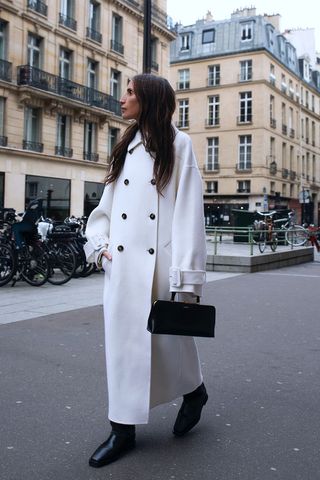 riding boot outfit shown in a photo of a woman in the street wearing a long white coat over a black dress with black riding boots and a black handbag