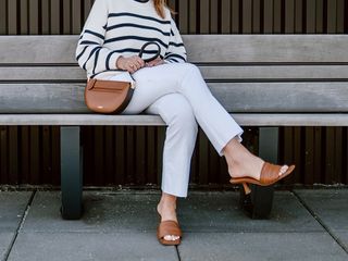 a woman sits on a bench while wearing white jeans and a white and black striped sweater with a brown crossbody leather bag and brown heeled mules