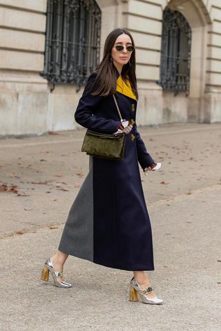 a photo of a woman's outfit with silver mary janes styled with a long navy coat and green shoulder bag and sunglasses
