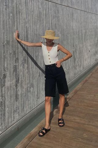 what to pack for cancun displayed on a woman wearing denim shorts with a white waistcoat, black sandals, and a woven hat