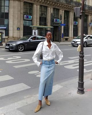 Sylvie wears a white button-down shirt, denim maxi skirt, beige suede mules, and a black bag.