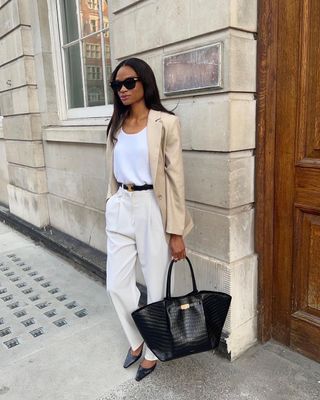 A woman wears a beige blazer, white tank top, black belt, ivory trousers, black flats, and a black tote bag.