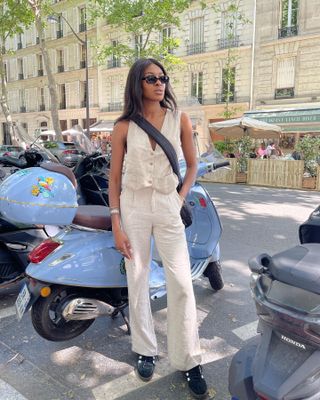 A woman wears a beige vest, beige trousers, black adidas sneakers, and a black crossbody bag.