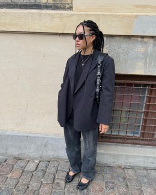 A woman wears an oversize black blazer, black jeans, maison margiela black flats, a grommet black bag, and black sunglasses.