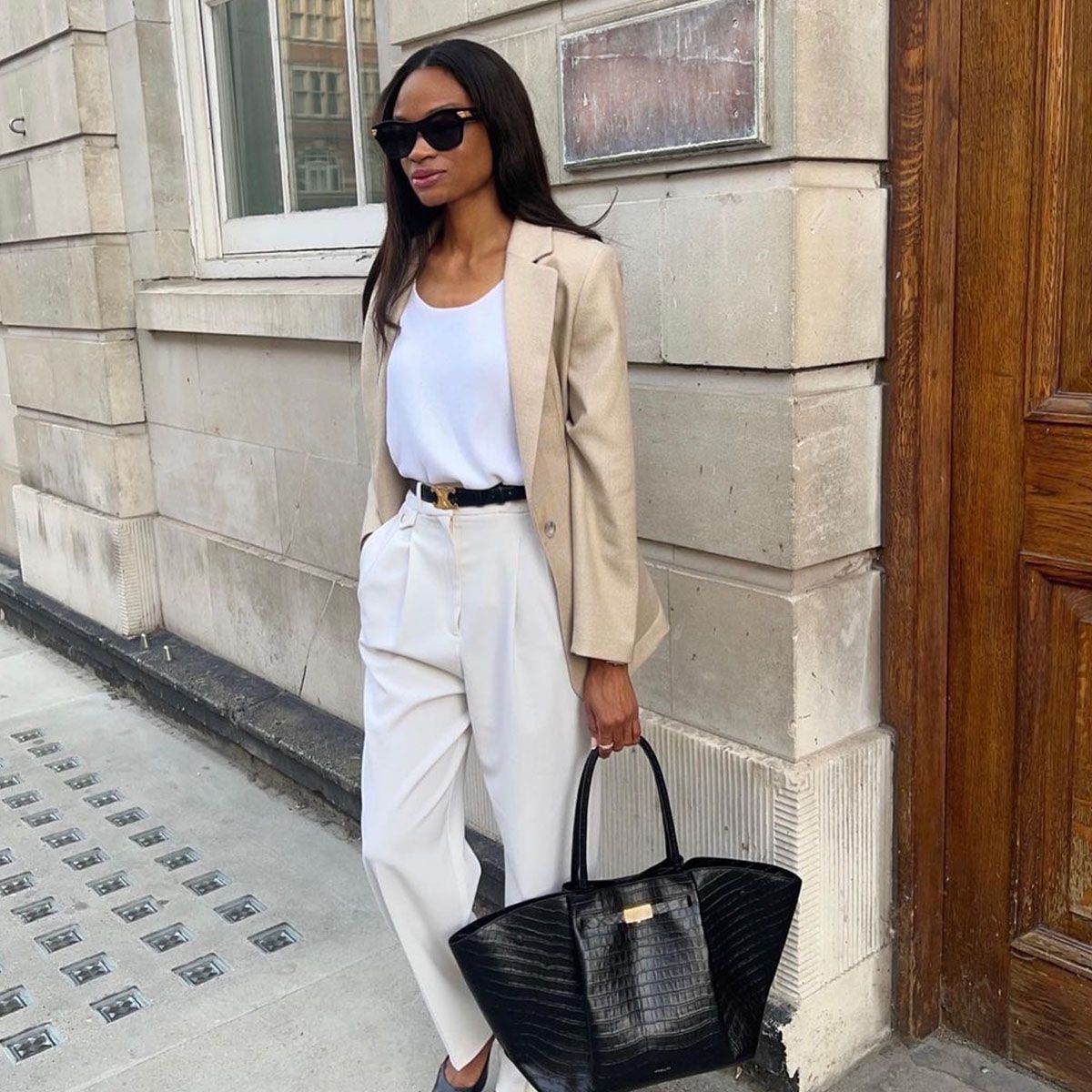 One woman wears a beige blazer, white tank top, black belt, ivory trousers and black tote bag.