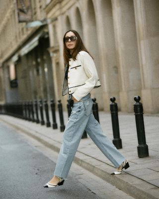 A woman wears a black and white jacket, blue jeans, and black and white heeled ballet pumps.