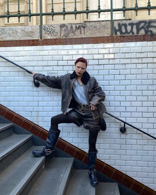 A woman wears a brown leather jacket with fur trim, a printed vest, a black belt, black leather shorts, black tights, and black knee-high engineer boots.